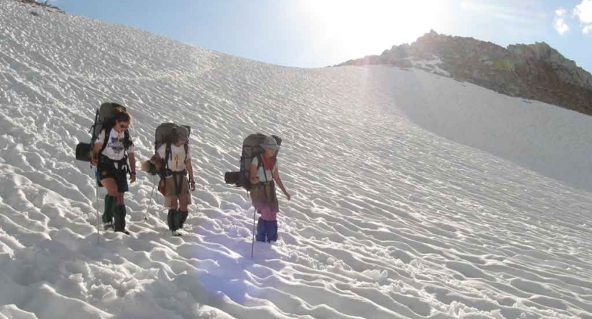 three people wearing backpacks hike down a snowy slope on an outward bound course